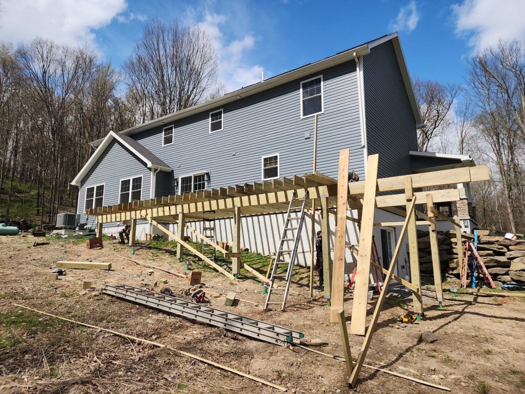 Early framing stage of a long deck expansion, showing pressure-treated floor joists and support beams being installed along the back of a house.